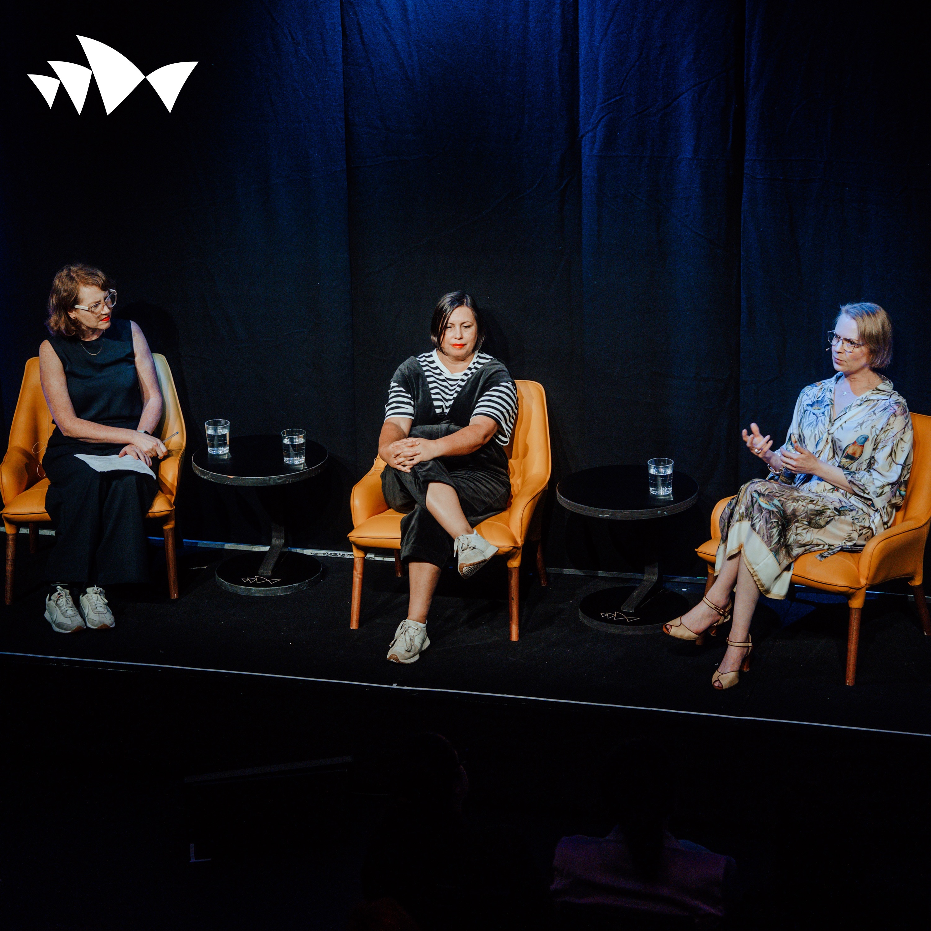 Three panelists sit in orange chairs on a dark stage, with small tables and water glasses during a discussion.