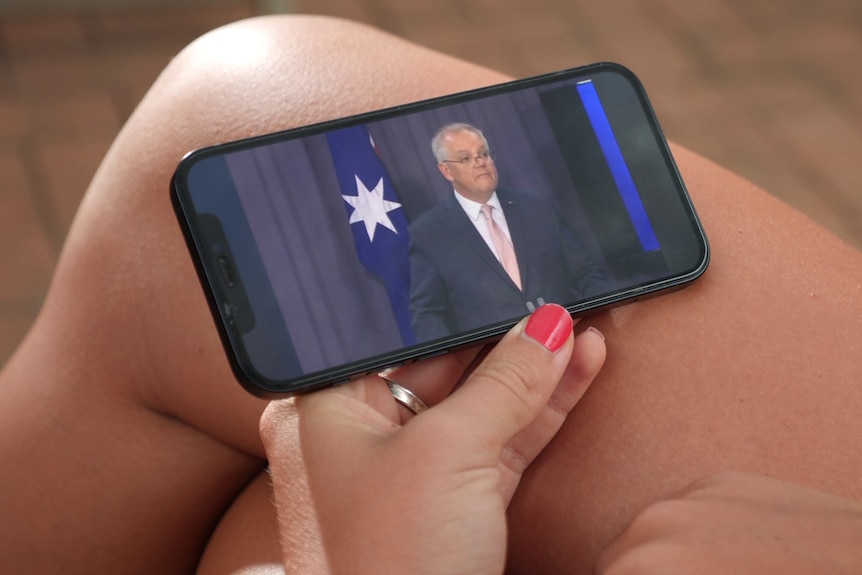 A young woman holding a phone, the phone playing a press conference featuring Prime Minister Scott Morrison