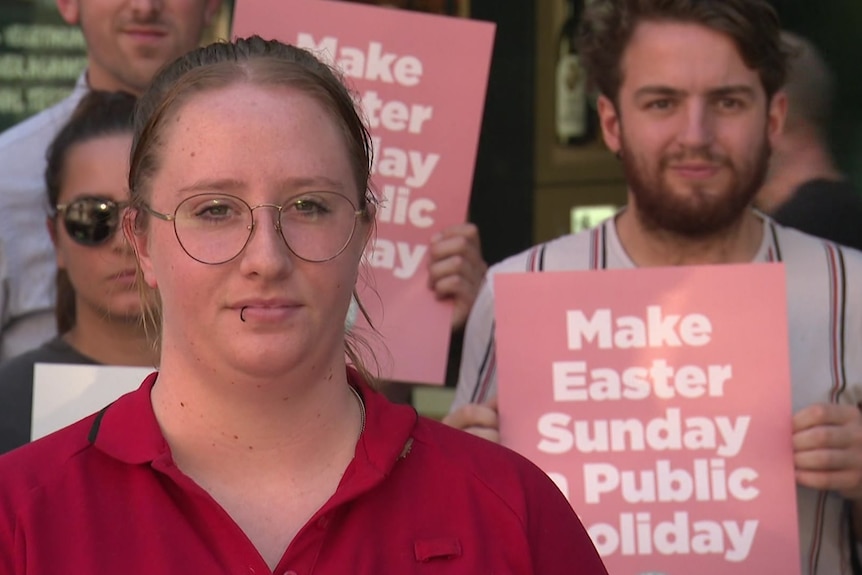 A woman wearing a red top and round glasses stands in front of people holding pink signs