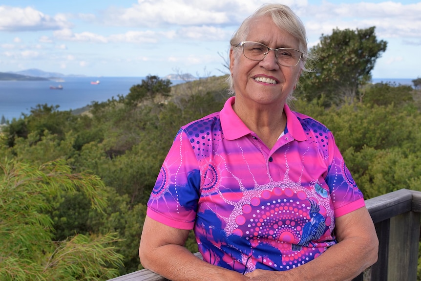 Woman wearing glasses smiles at camera in front of lush green bush and ocean