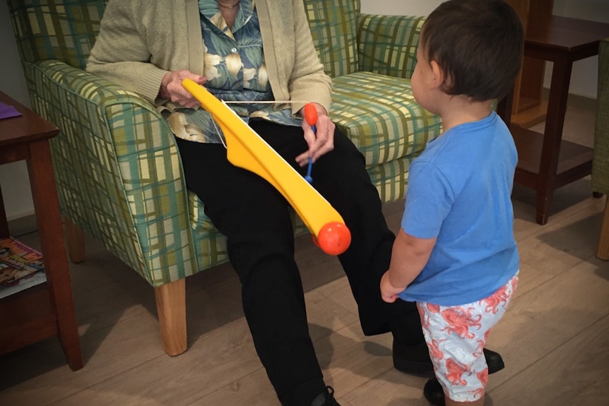 A toddler stands next to an elderly resident with dementia during a playgroup session.