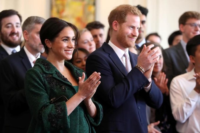 Meghan, left, wears a green dress and matching coat and claps next to Harry, right, who wears a navy suit. Both are smiling.