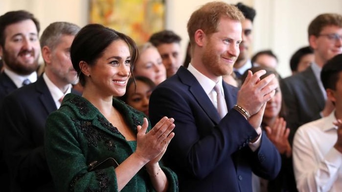 Meghan, left, wears a green dress and matching coat and claps next to Harry, right, who wears a navy suit. Both are smiling.