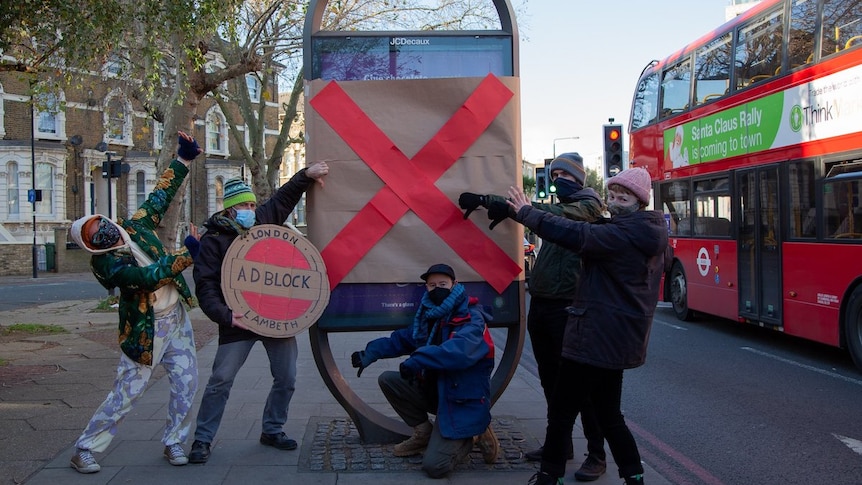 A group  of activists in facemasks gather around a billboard they have covered up, holding an Adblock London sign