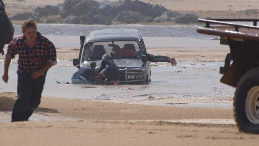 Guy running towards camera with a 4WD sinking in water with people tending to it in the background.