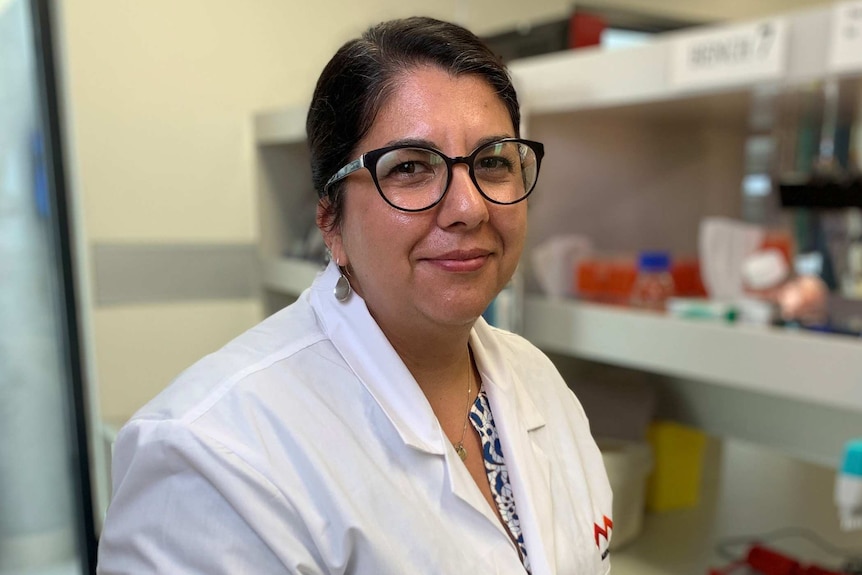 A woman wearing a white lab coat and glasses stands in a lab posing for a photo.