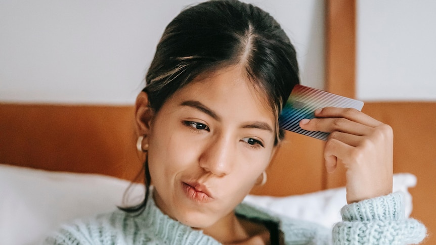 A stock image of a Woman in bed holding a credit card, looking pensive.