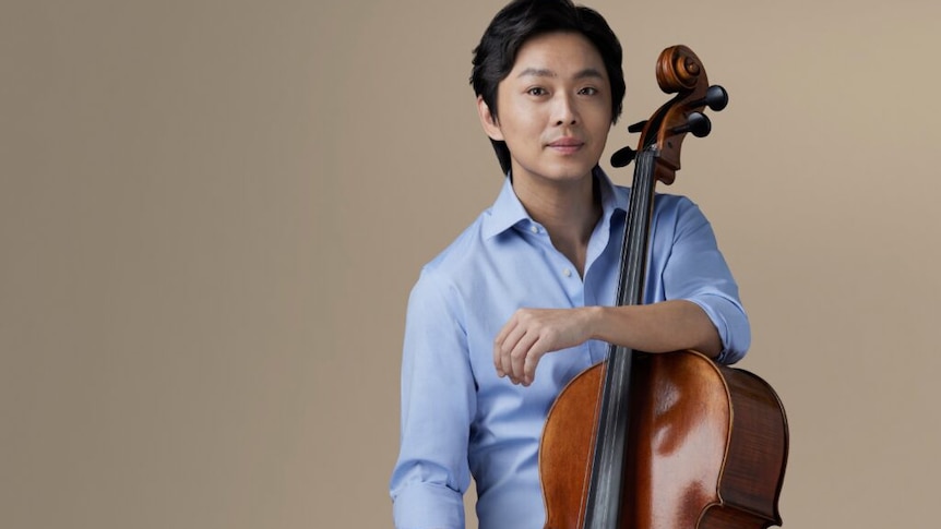 Li-Wei Qin in a blue shirt and white pants, his arm around the neck of his cello.
