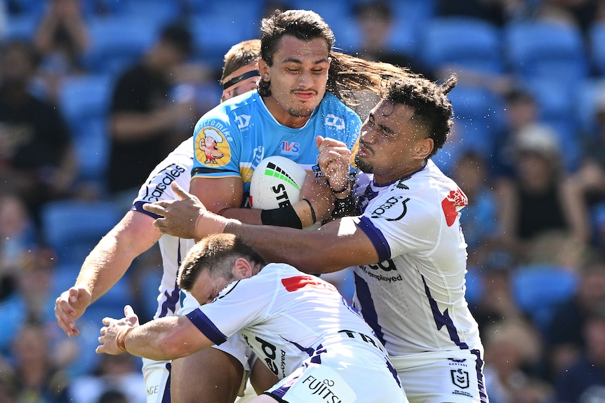 A Gold Coast Titans NRL player holds the ball as he is tackled by Melbourne Storm opponents.