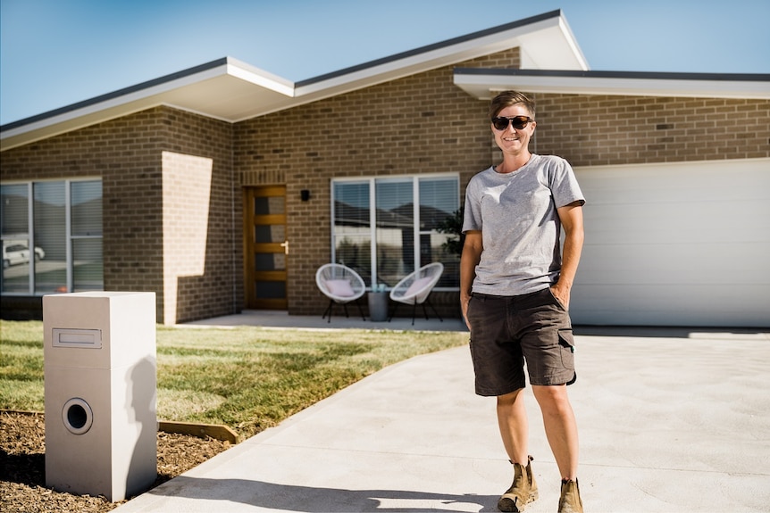 A woman wearing a t-shirt, shorts and work boots smiling and standing on the driveway of a house