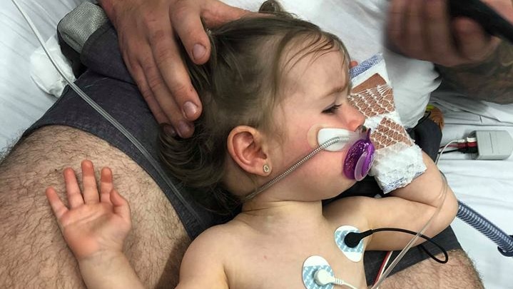 Wynter lies on her father's lap. She has monitors attached to her chest and stomach and a breathing mask below her nose.