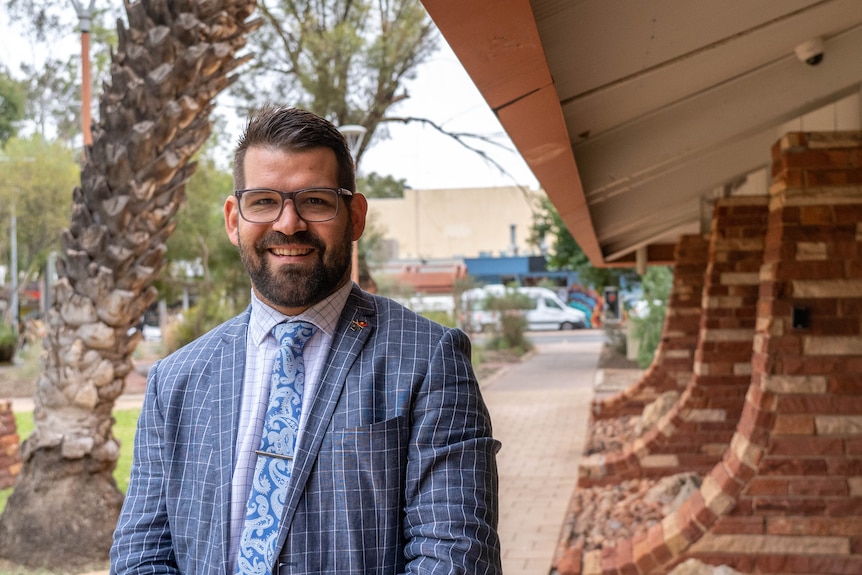 A smiling man with cropped dark hair, beard, wears glasses, blue check shoes and tie. Stands near brick wall and palm tree.
