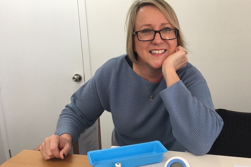 A fair-haired, middle aged woman wearing dark-rimmed glasses and a grey jumper with vaccine implements in front of her