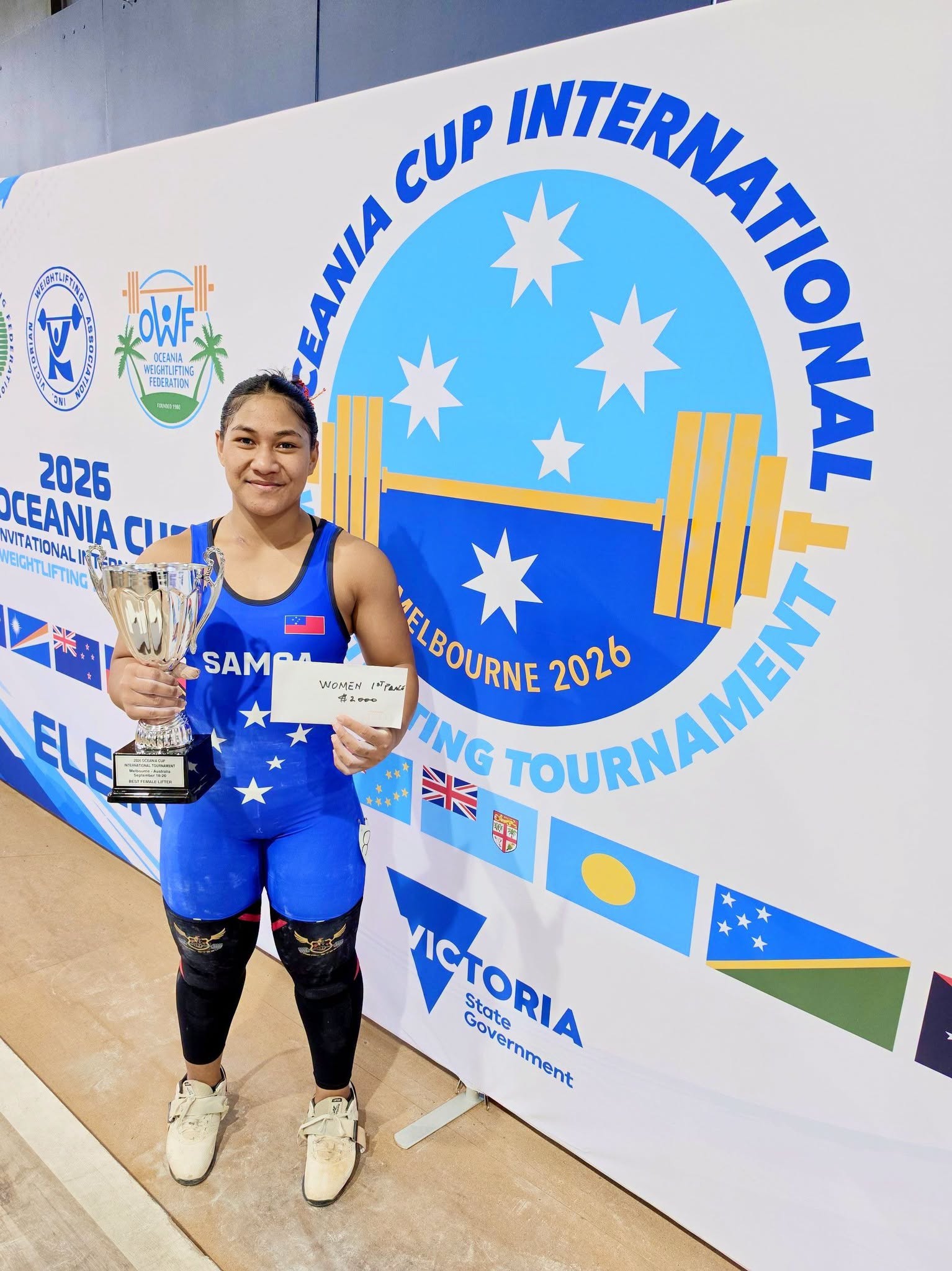 A young Samoan girl wearing a blue weightlifters tights holds a trophy in one hand and a cheque in the other hand and smiles.