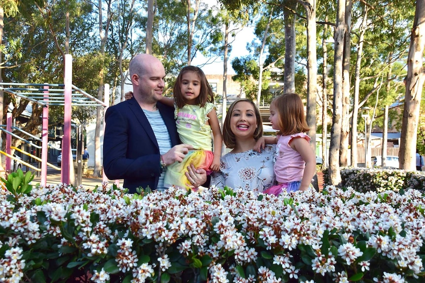 A man and woman holding two young girls standing behind a flowering bush, illustrating a happy family after a stillbirth.