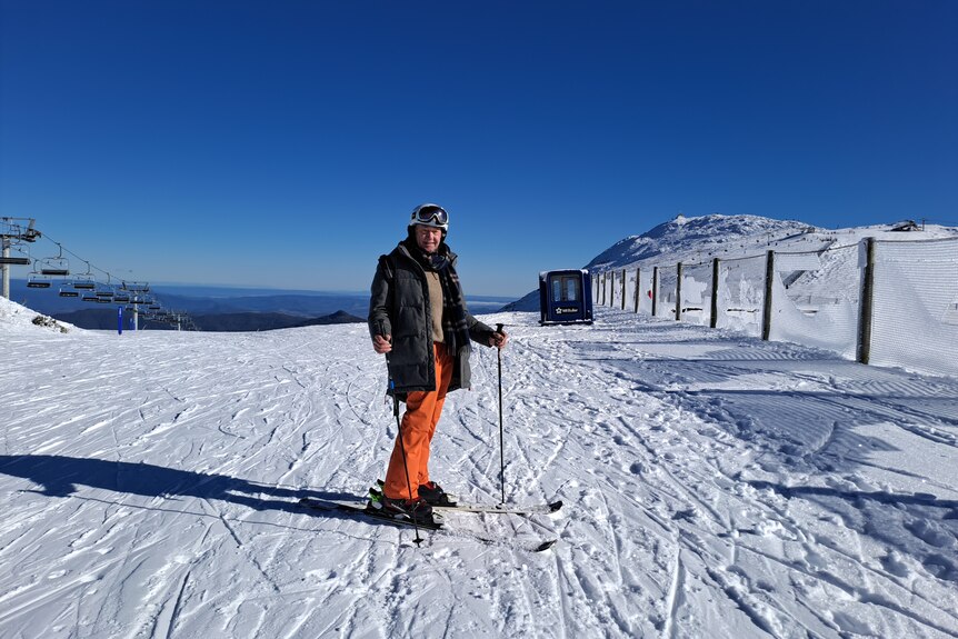 A man wearing skis stands facing the camera, he is wearing orange ski pants and a dark green jacket.