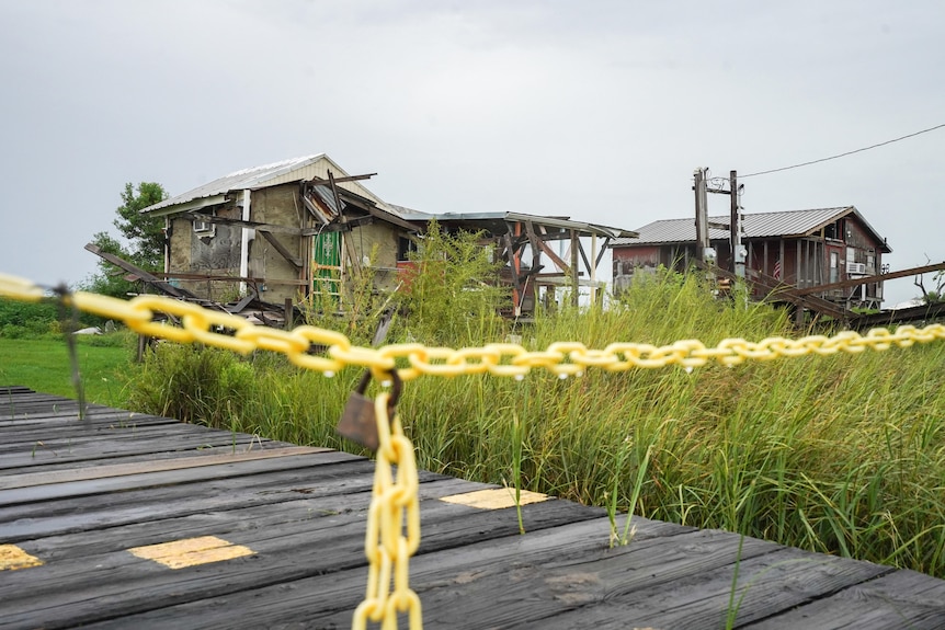 a damaged home behind a yellow chain with a lock and weeds growing behind it 