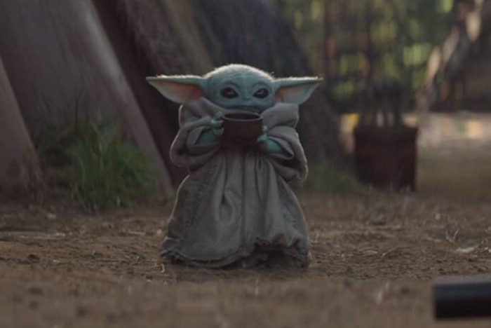 The Mandalorian character Baby Yoda holds a brown soup bowl, wearing a brown robe.