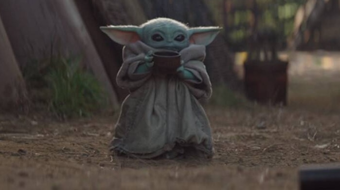 The Mandalorian character Baby Yoda holds a brown soup bowl, wearing a brown robe.