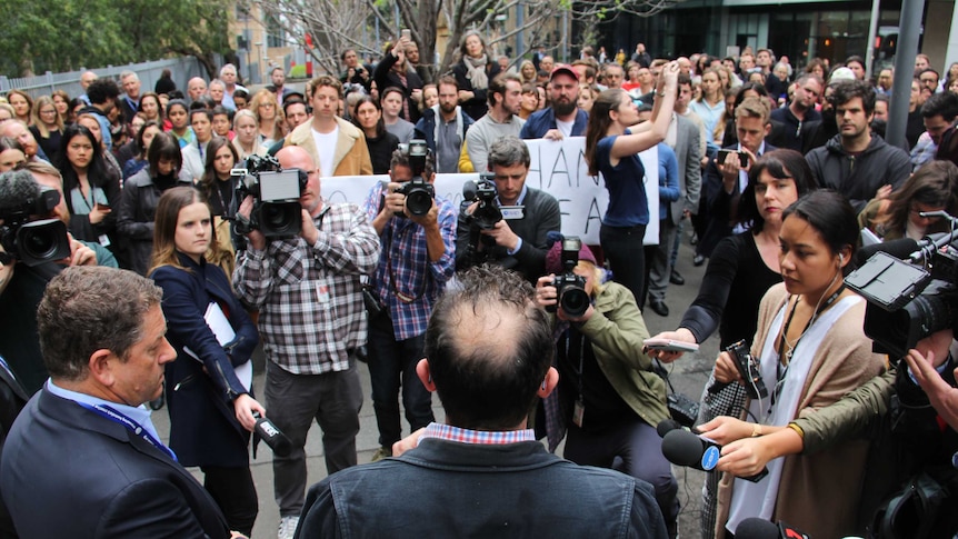A crowd of people, some with cameras, microphones, phones and placards, gather around Miskelly