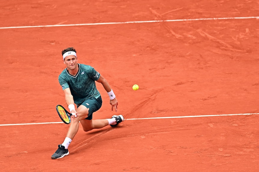 A young tennis player with a headband on his head slides across the red clay to hit a forehand return.