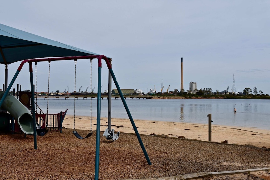 A children's playground with no one using the equipment, in the background across a the water is a lead smelter.