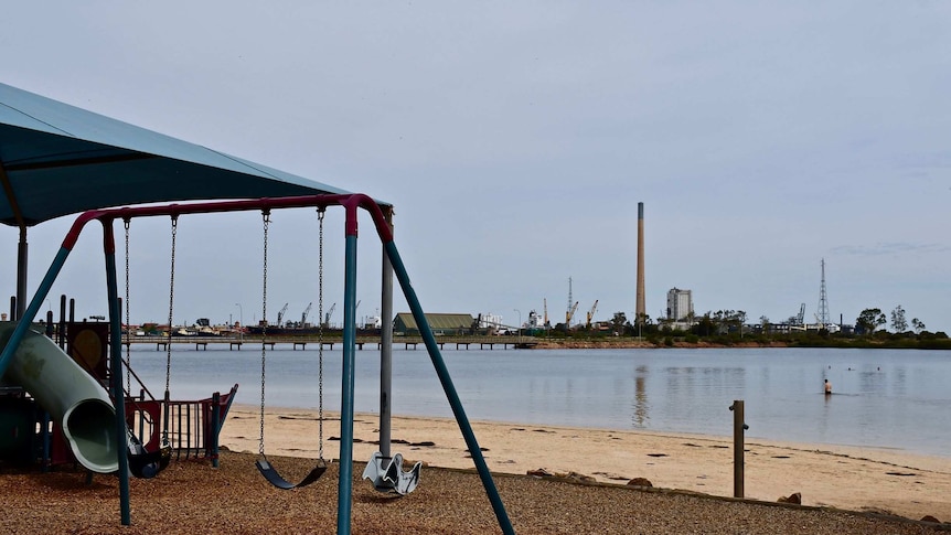 A children's playground with no one using the equipment, in the background across a the water is a lead smelter.