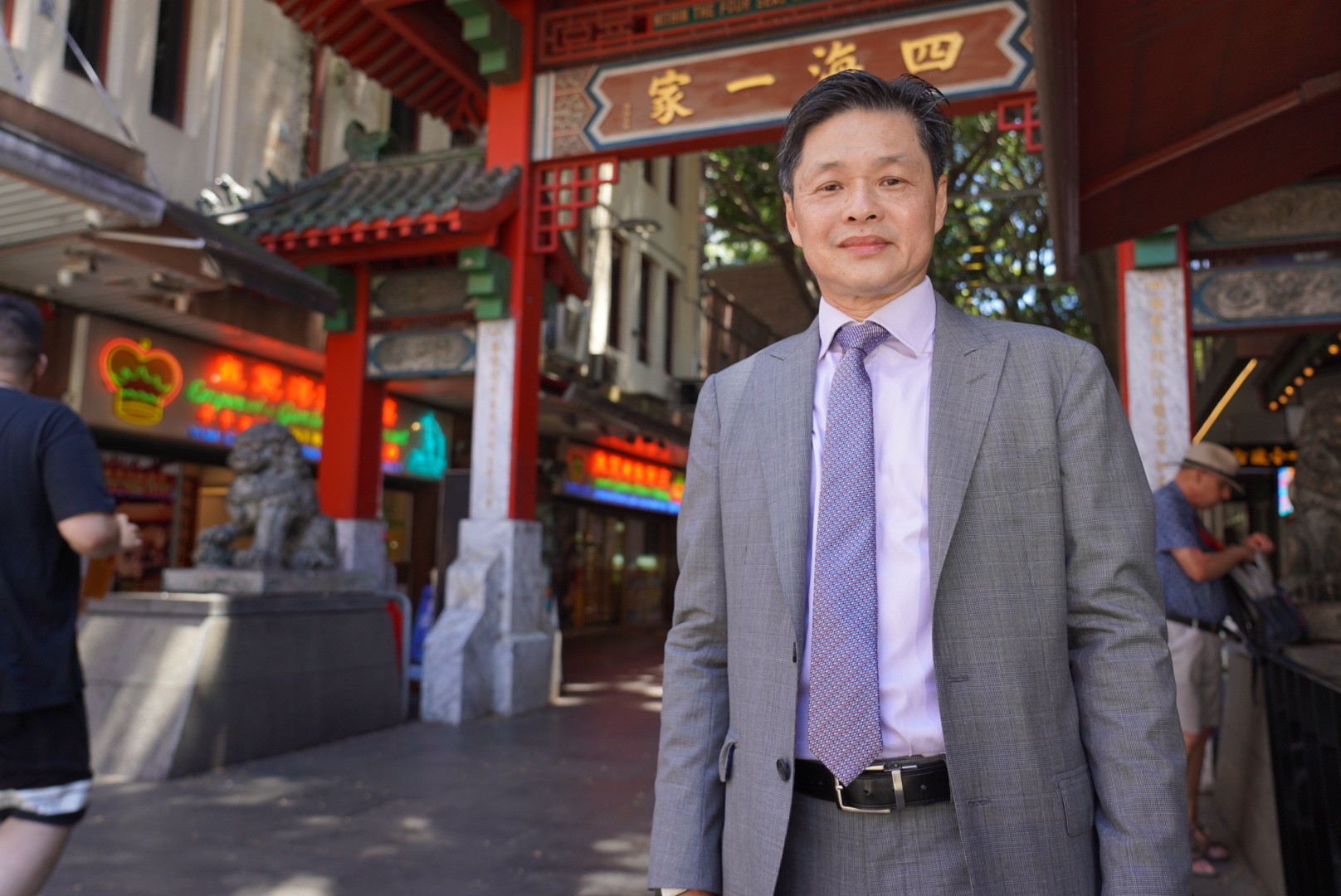 A man wearing a grey suit and lavender tie and shirt stands in front of a Chinese paifang and restaurants.