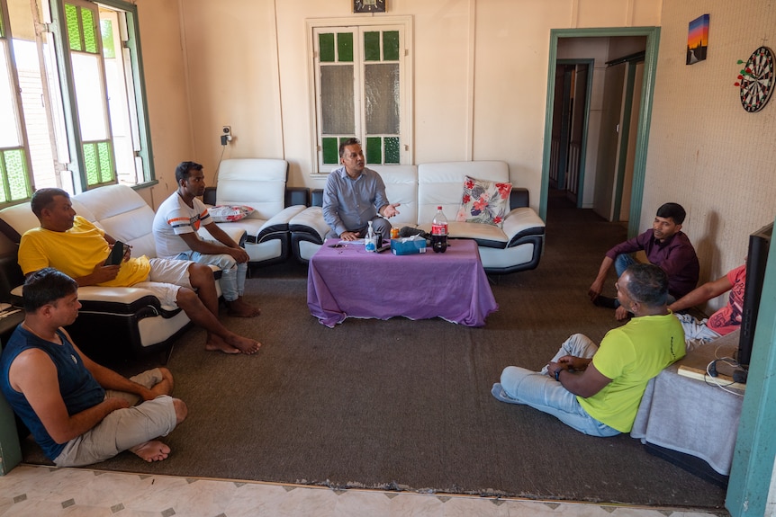 A group of men from Bangladesh sit in a large living room in a rundown sharehouse.