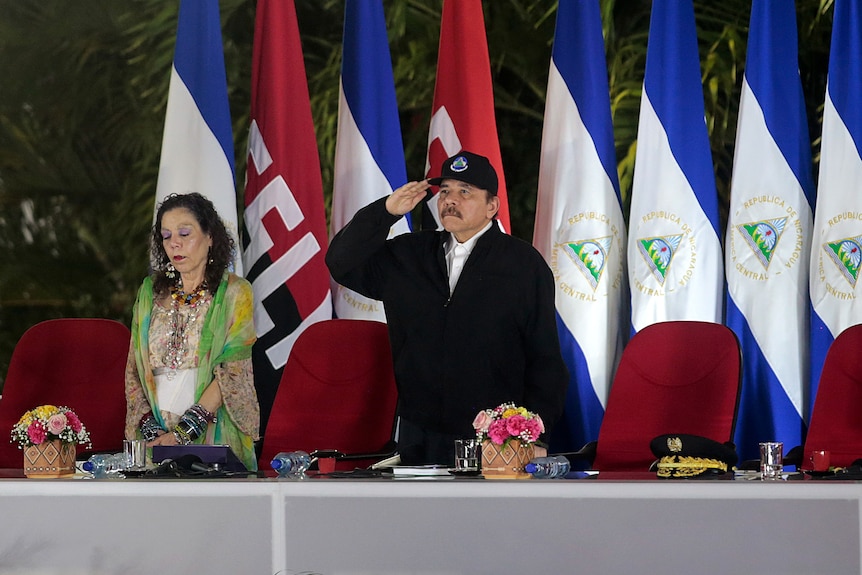Nicaragua's' President Daniel Ortega stands and salutes in a black cap in front of a Nicaraguan flag
