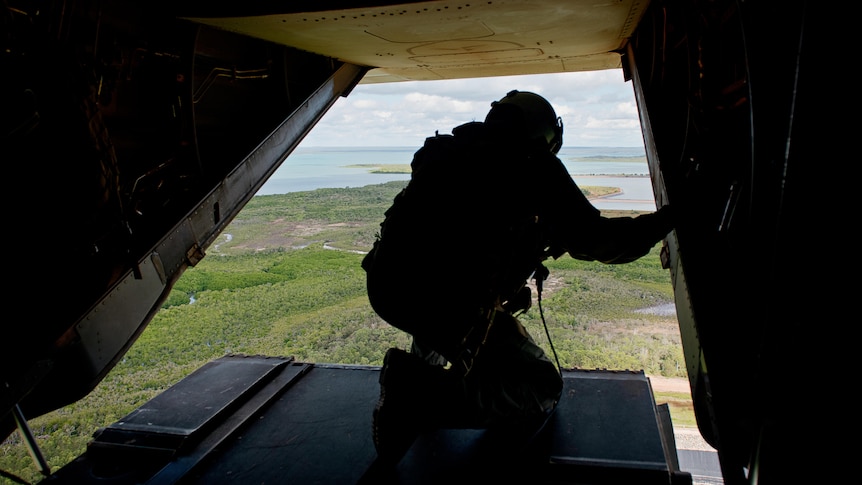 Looking out at a soldier crouched on the loading tray of a military aircraft flying over green landscape, water in the distance