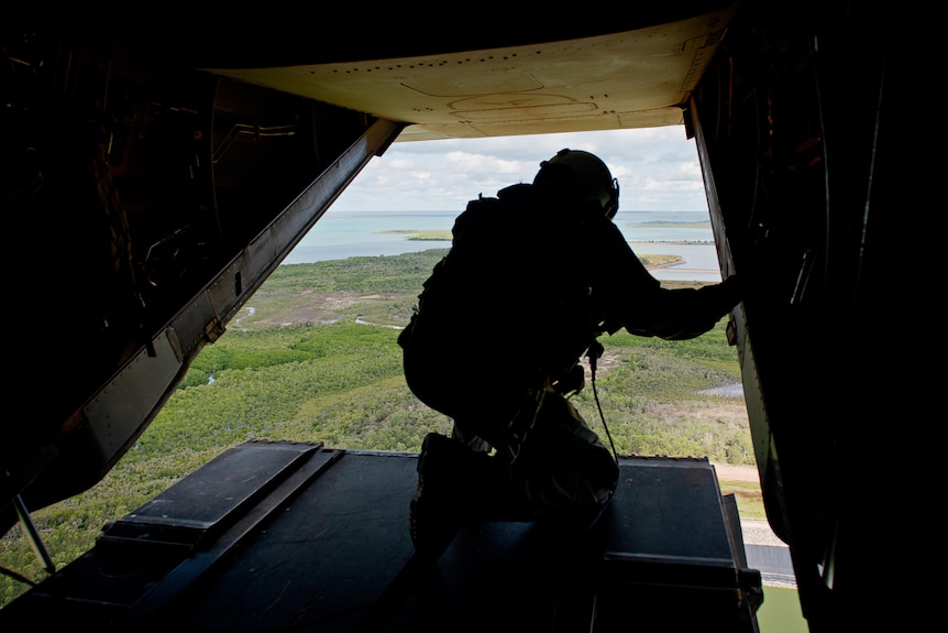 Looking at a soldier crouched on the cargo bed of a military aircraft flying over a green landscape, water in the distance