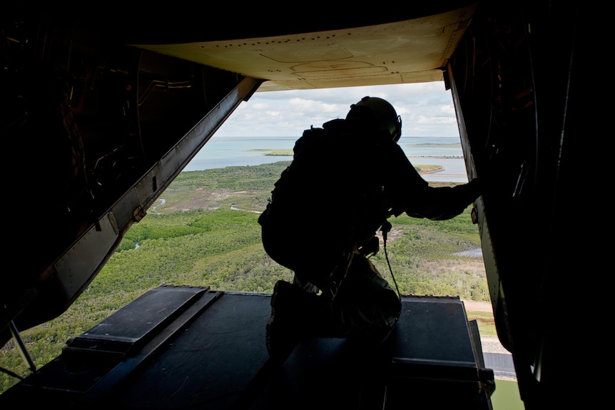 Looking out at a soldier crouched on the loading tray of a military aircraft flying over green landscape, water in the distance