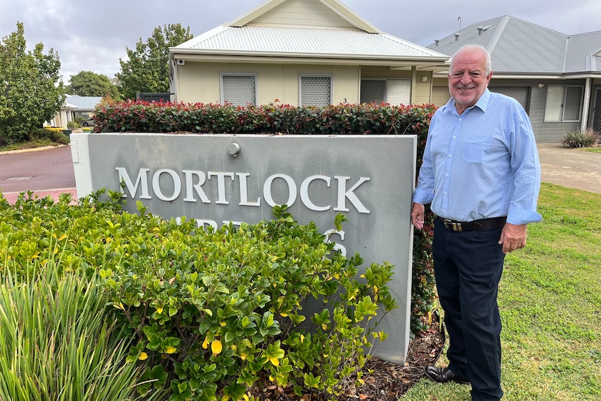 A man wearing a blue business shirt and dark blue dress pants is similing next to a grey sign that says 'Mortlock'