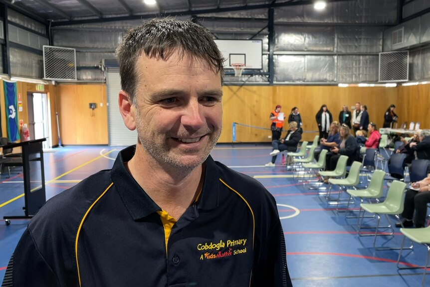 A man wearing a navy and yellow shirt smiles near the camera, inside a school gym.