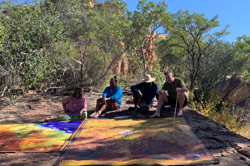four people sitting down looking over a large map on a flat rock surface in the bush