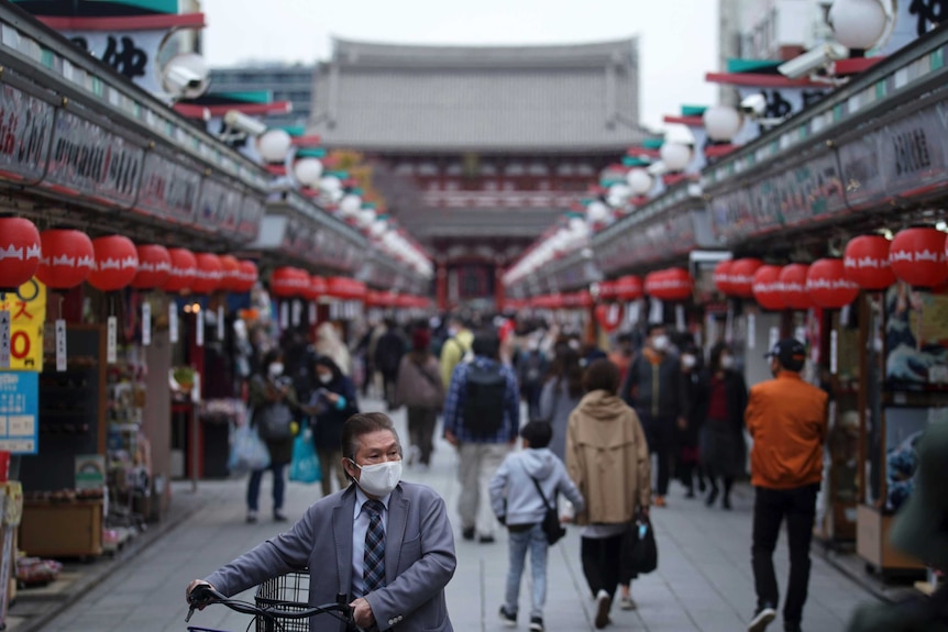 A person wearing a mask and holding a bike stands in front of people walking through a mall in Japan.