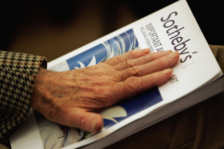 An older person's hand rests on a Sotheby's catalog.