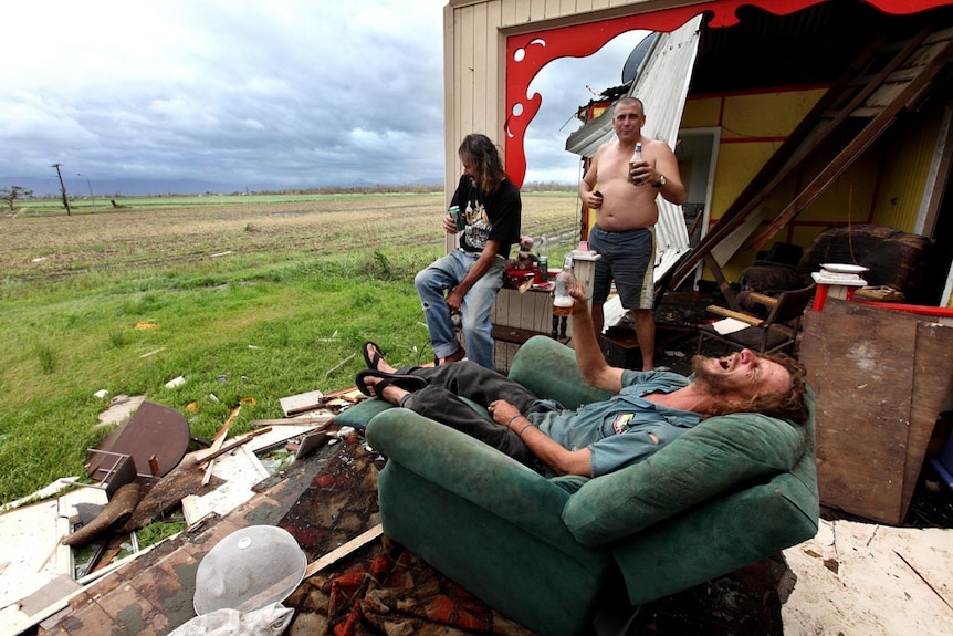 Three men, one stands, one rests on shelf, another lies on armchair, all raise their beers in ruined house looking onto field.