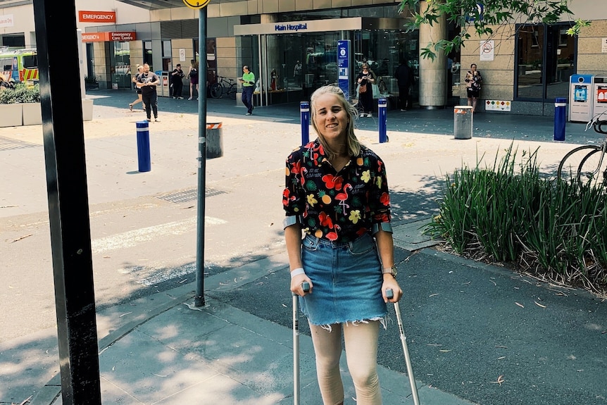 A young woman wearing a black and red florat shirt, denim skirt and converse stands with crutches outside a hospital.