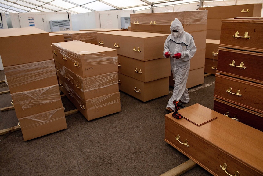 A person in HAZMAT gear is surrounded by coffins as they walk through a makeshift morgue.