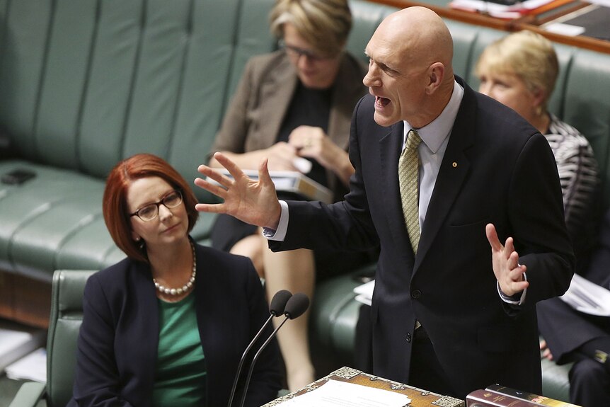Politician Peter Garrett wears a suit standing up speaking during question time, seated is politician Julia Gillard