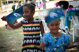 Two smiling young Indigenous girls in colourful dresses happily wave small blue, aqua, black and white flags