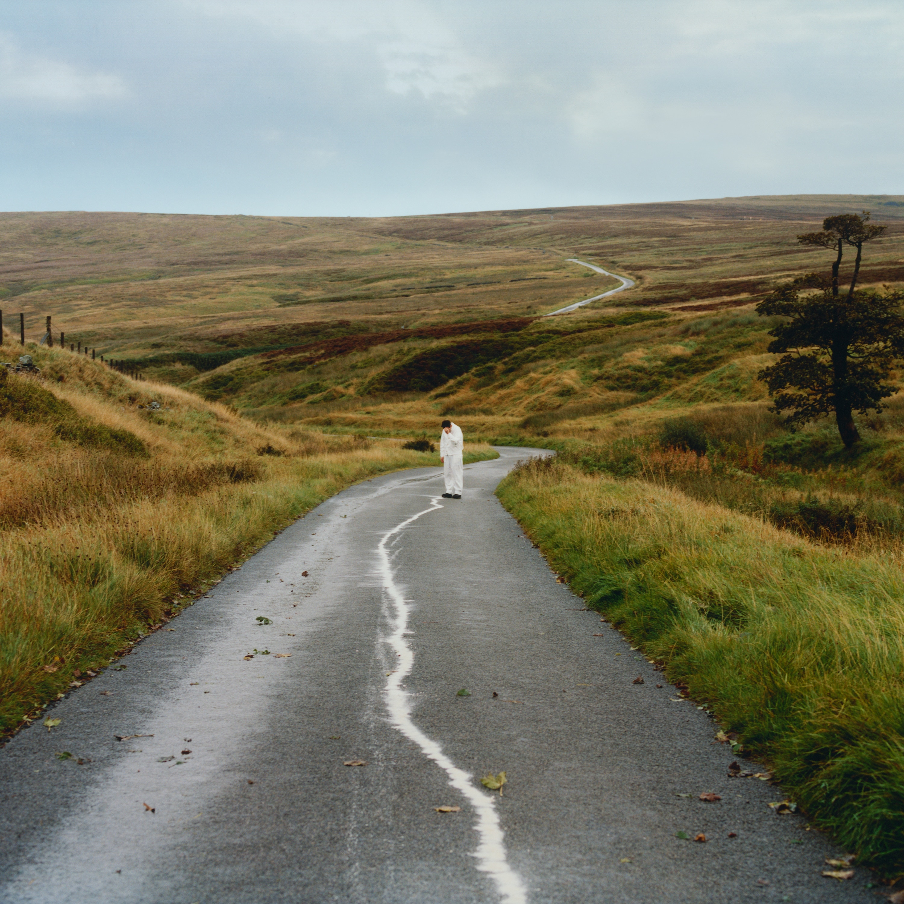 Man in all-white coveralls stands in the distance on a country road with rolling brown green hills and lone tree