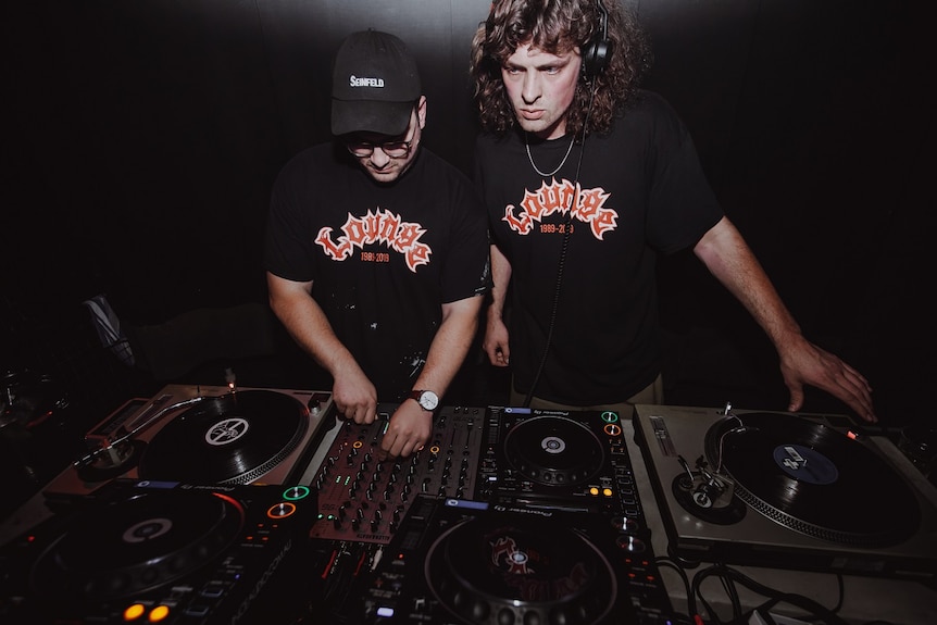 Two men in black t-shirts stand behind turntables in a nightclub.
