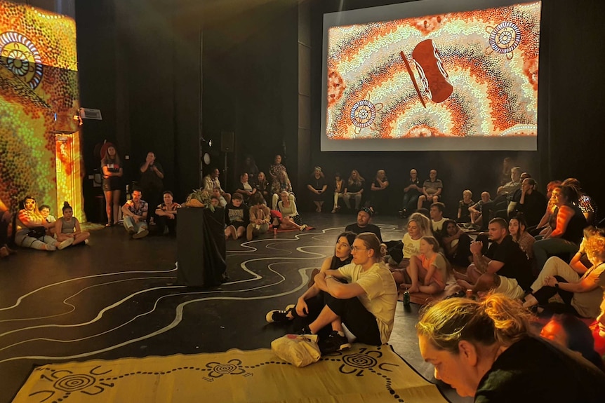 Large screens projecting indigenous art work on the right and left, a crowd of 50 people on floor looking to left of photograph