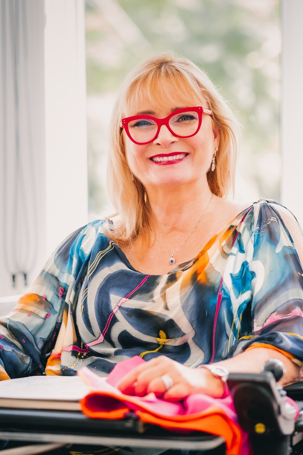 A woman with bright red glasses sits at a desk holding a fabric swatch.