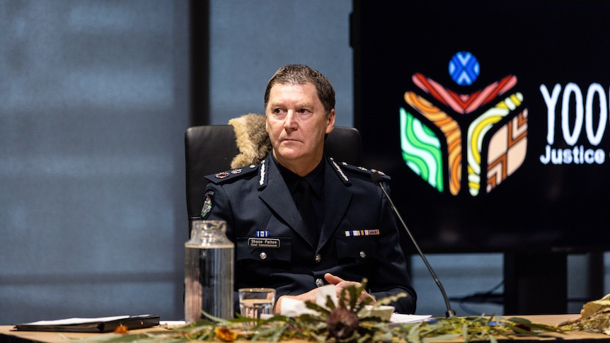 Shane Patton is in his navy blue full police chief uniform as he sits looking seriously at a table adorned with native leaves.