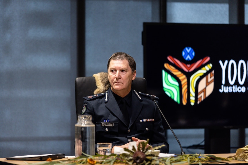 Shane Patton is in his navy blue full police chief uniform as he sits looking seriously at a table adorned with native leaves.