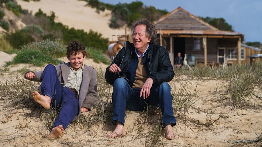 Geoffrey Rush sits next to a boy on the sand during the filming of Storm Boy.